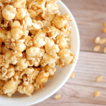 white bowl filled with gooey caramel popcorn and popcorn kernels scattered on the table next to the bowl