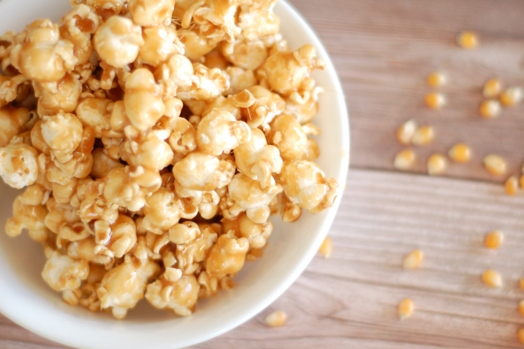 white bowl filled with gooey caramel popcorn and popcorn kernels scattered on the table next to the bowl