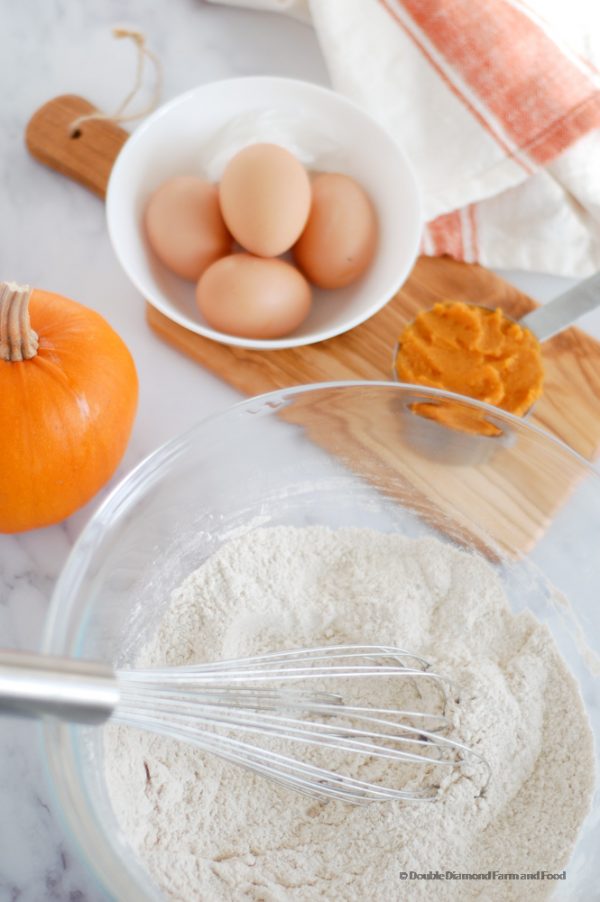 A glass bowl with the flour and spice. A white bowl with fresh farm eggs and a measuring cup with pumpkin puree.